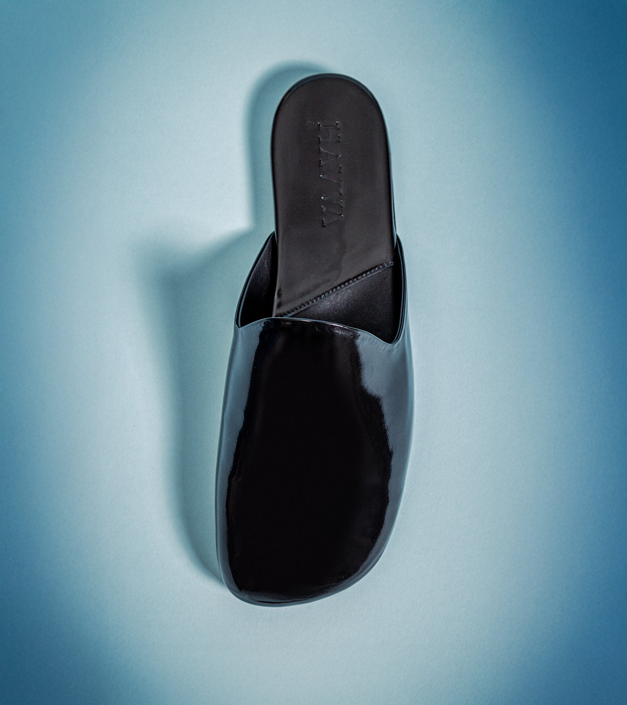A single black patent leather mule sandal with a square toe, shown from a top-down perspective on a blue gradient background.